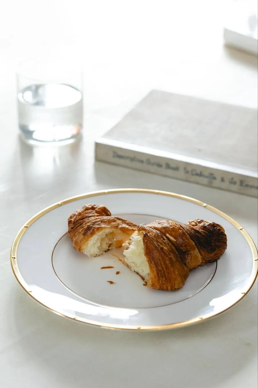 クロワッサンが半分に割られて、金色の縁取りのある白い皿に置かれています。グラスに入った水と、本が背景に写っています。