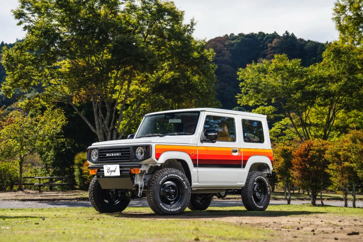 白いスズキジムニーが芝生の上に駐車されています。車体側面にはオレンジ、黄色、赤色のストライプが入っています。