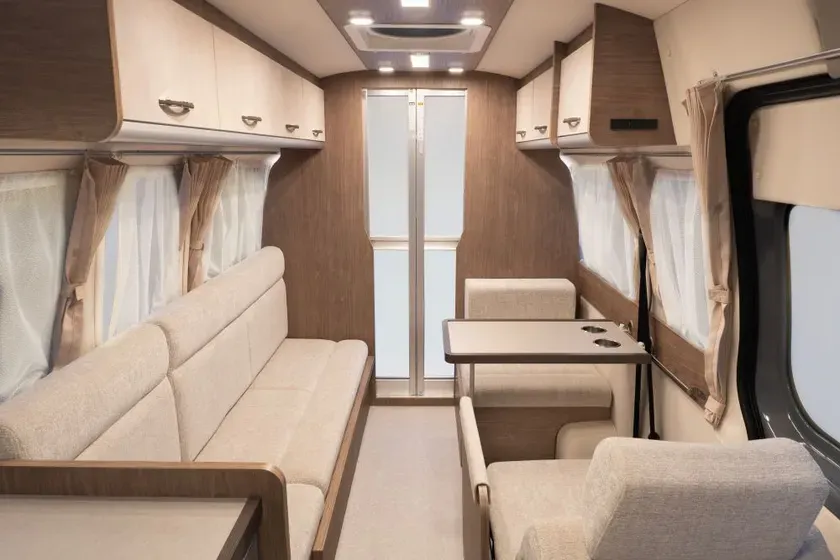 キャンピングカーのインテリア。木製のキャビネット、ソファ、テーブル、窓、カーテンが写っています。