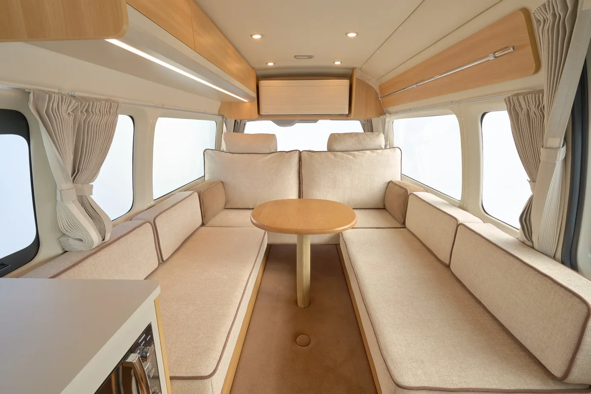 キャンピングカーのインテリア。ベージュのソファと丸いテーブルがあり、窓から自然光が入る。