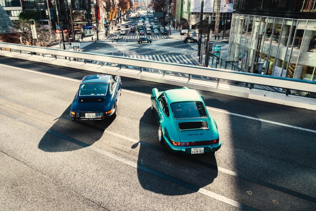 高架道路を走行する2台のヴィンテージポルシェ（青とティールグリーン）を上空から捉えた写真です。下には多くの車が行き交う賑やかな都市の交差点が広がっており、建物や横断歩道が見えます。日差しが強く、車や欄干の影が長く伸びています。