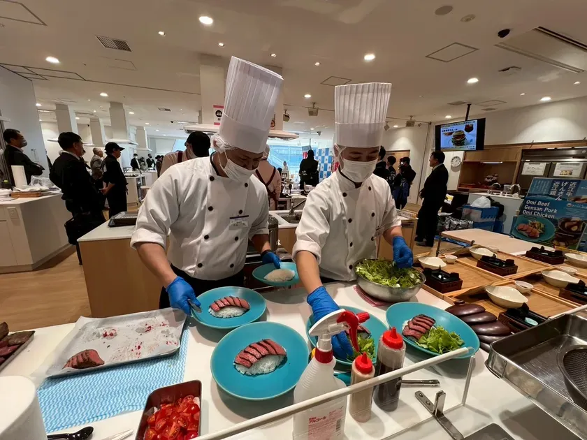 2人のシェフが料理を作っている様子。皿に盛り付けられた肉とご飯、サラダが見える。周りには見学者がいる。