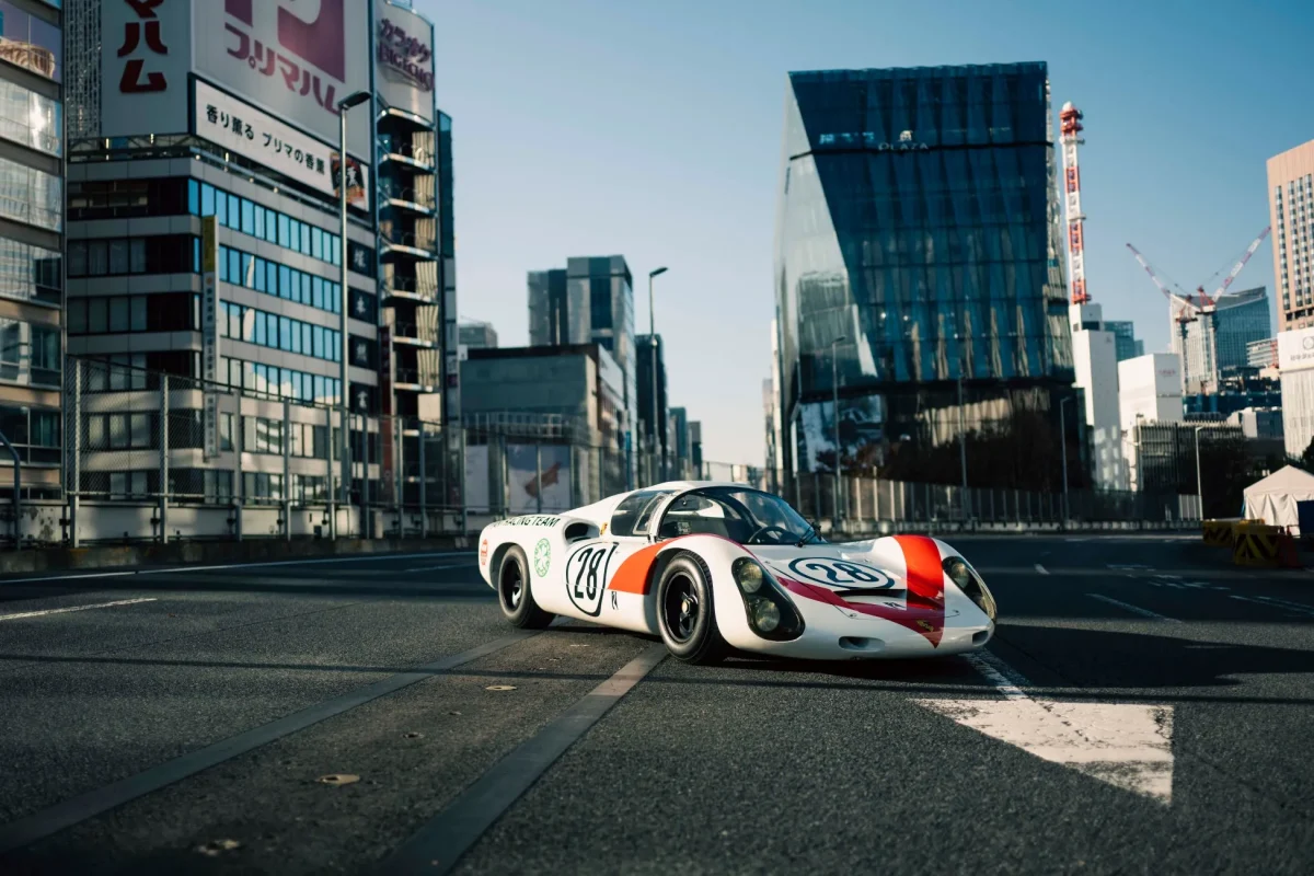都市の道路に停まっているクラシックカーの写真。白と赤のレーシングカーで、番号28が側面に表示されています。