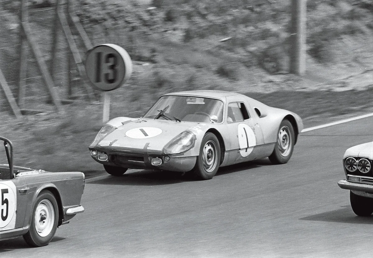 レース中のヴィンテージカーの白黒写真。ナンバー1の車がリードしており、ナンバー13の標識が背景に見える。