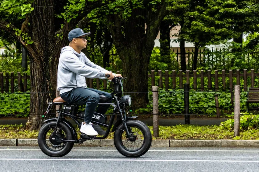電動バイクに乗る男性の画像。男性はグレーのパーカーとキャップを着用し、サングラスを着用している。背景には木々や緑の植物が見える。