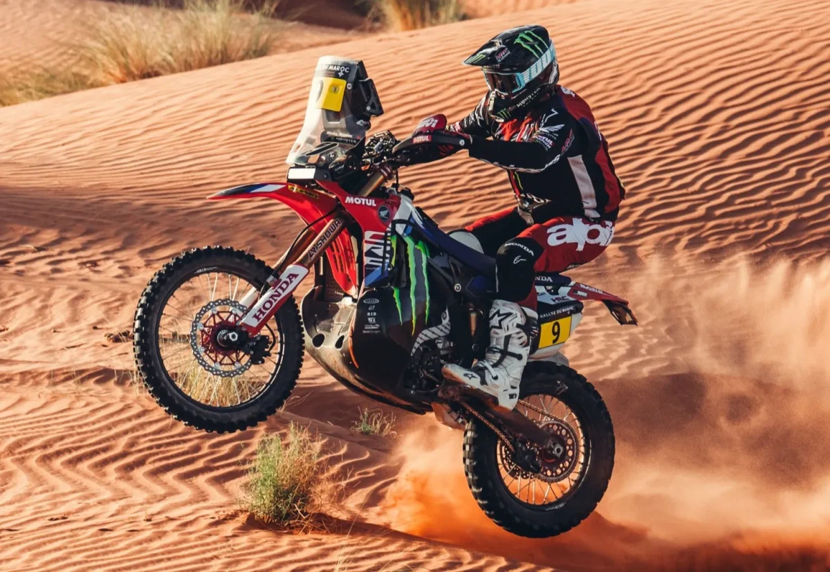 砂漠でバイクに乗っているレーサーの画像。バイクは空中にあり、ライダーはヘルメットをかぶっています。
